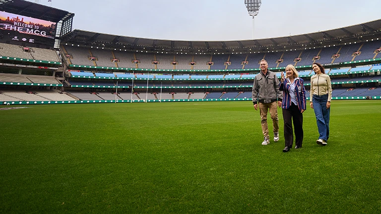 Group of 3 walks across the MCG turf during a stadium tour