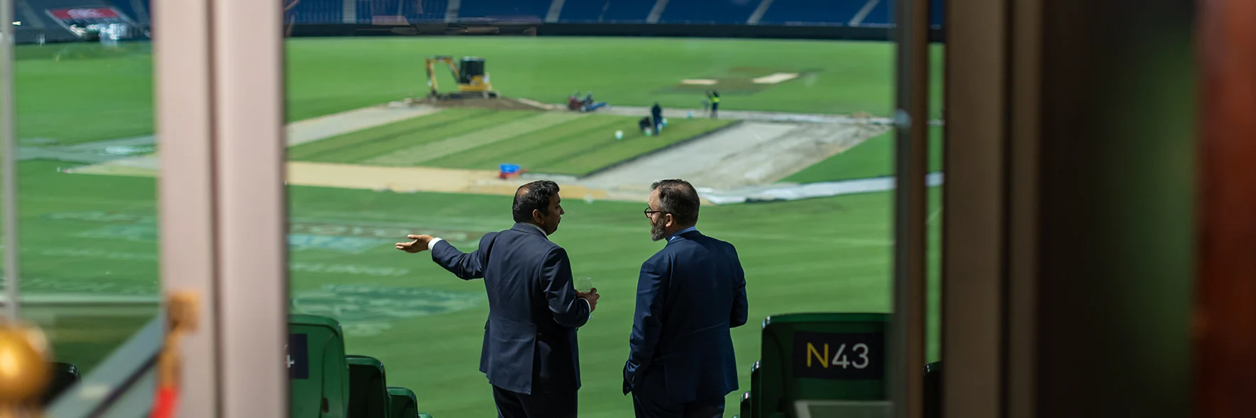 Two guests in suits looking out as drop-in wicket work takes place on the MCG arena