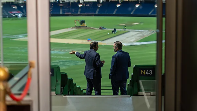 Two guests in suits looking out as drop-in wicket work takes place on the MCG arena