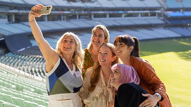 A group of five women taking a selfie together in the stands at the MCG
